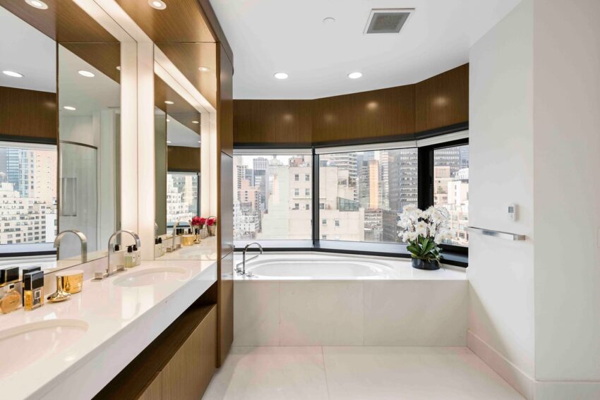 Modern bathroom with a large tub, dual sinks, mirrored cabinets, city skyline view, and decorative flowers.