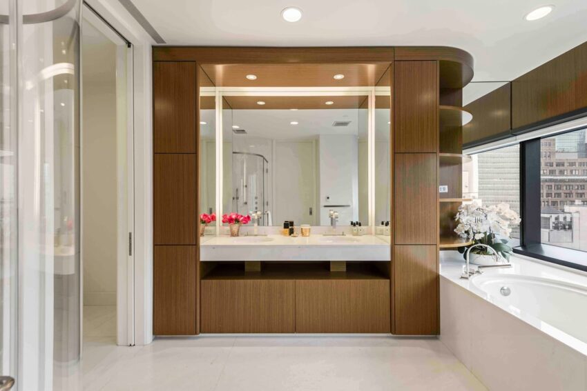 Modern bathroom with wooden vanity, large mirror, pink flowers, and a bathtub next to a window with city view.