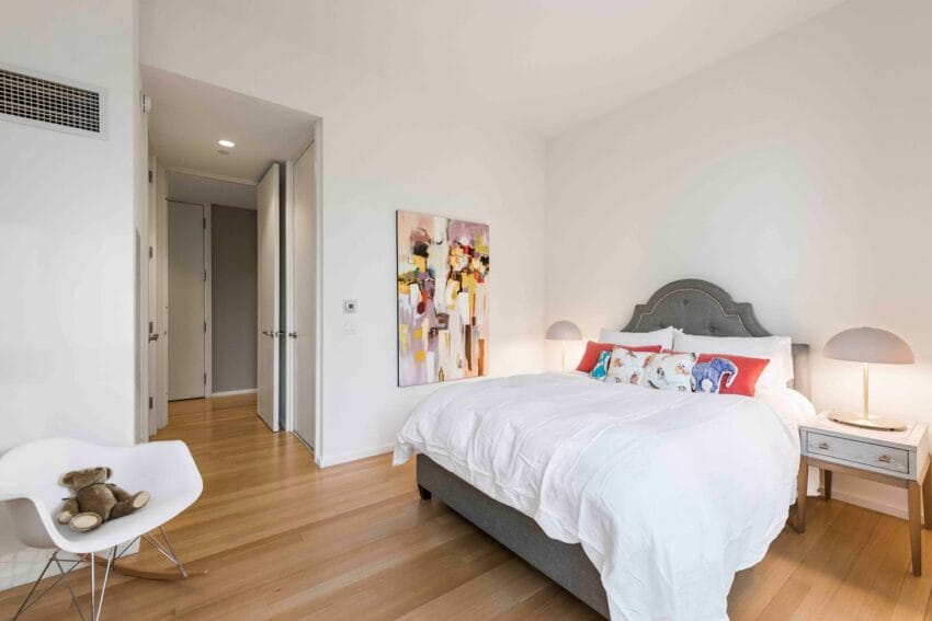 Modern bedroom with a large bed, decorative pillows, a colorful painting, wooden floor, and a small chair with a teddy bear.