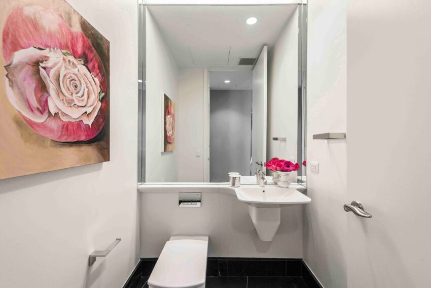 Small modern bathroom with pink rose painting, wall-mounted sink, toilet, and a bouquet of pink flowers on the counter.