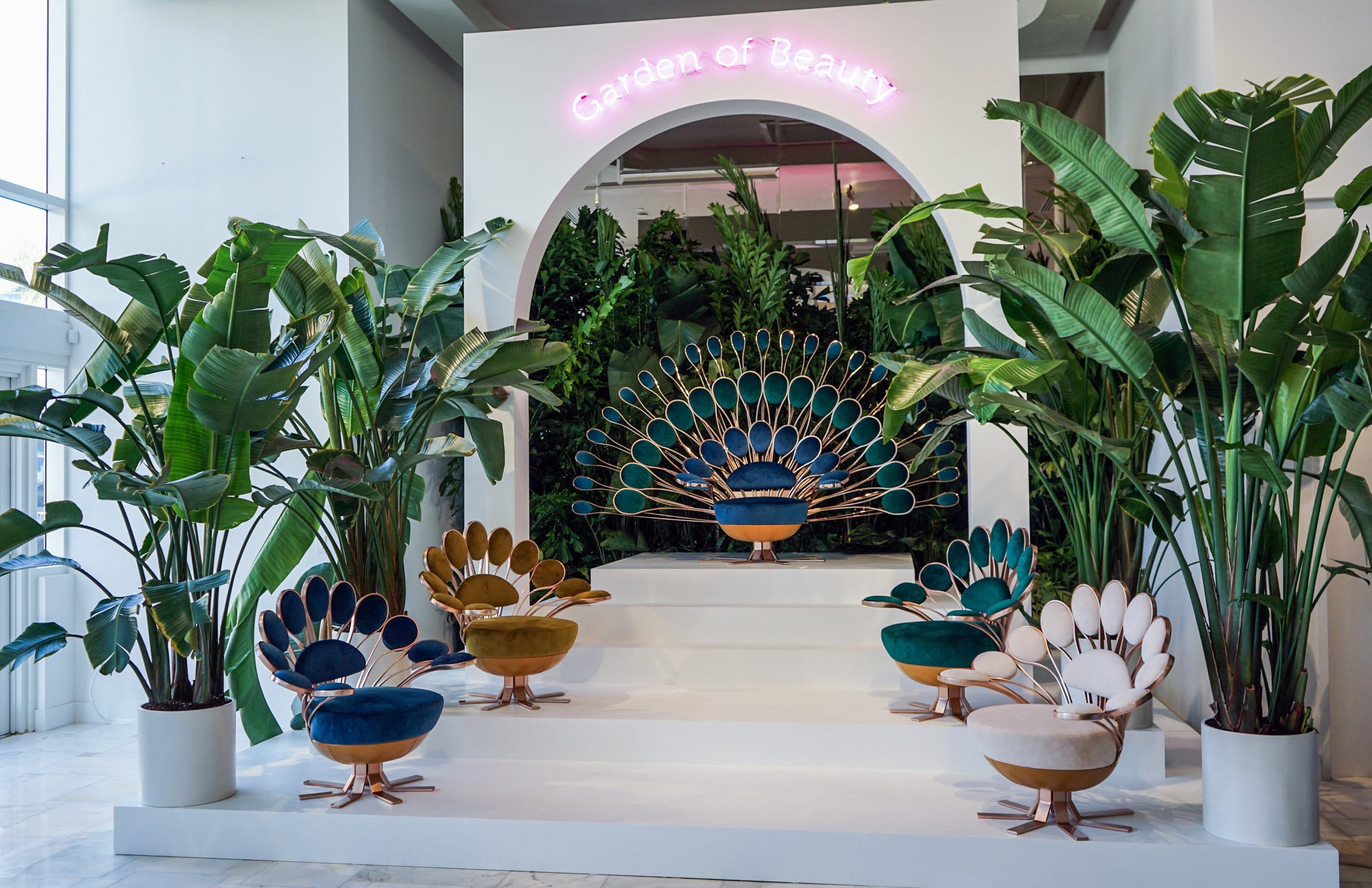 Peacock chairs on a white platform surrounded by potted plants under a neon garden of beauty sign.