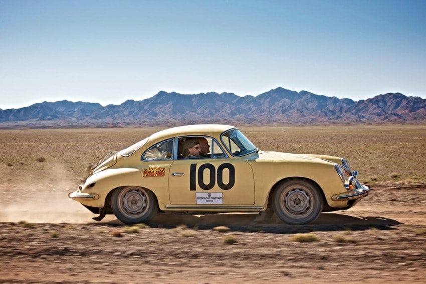 Vintage yellow race car with number 100 speeding across a desert landscape with distant mountain range in the background