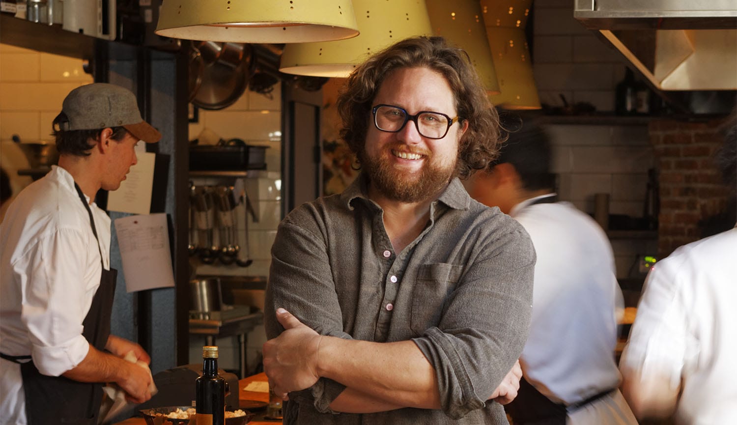 Smiling man with glasses standing in a busy restaurant kitchen with chefs working in the background.