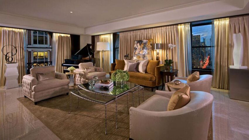 Elegant living room with plush sofas, glass coffee table, grand piano, and city view through large windows at dusk.