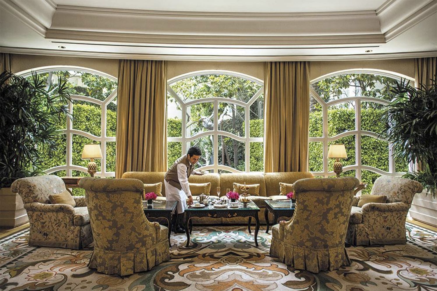 Luxurious hotel lounge with elegant furniture, large windows, lush plants, and a staff member serving tea.