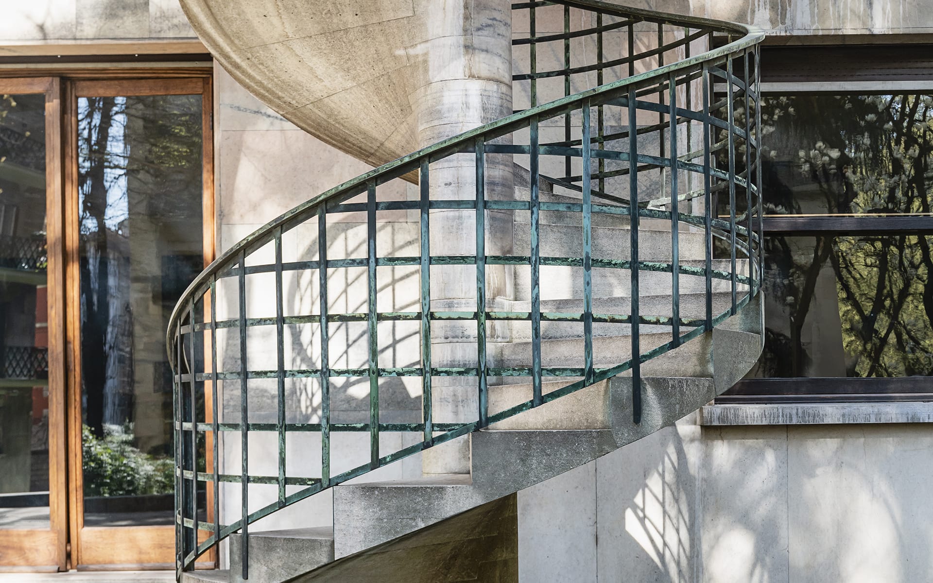 Spiral concrete staircase with green metal railing outside a building, with sunlight and shadows on the steps and wall.