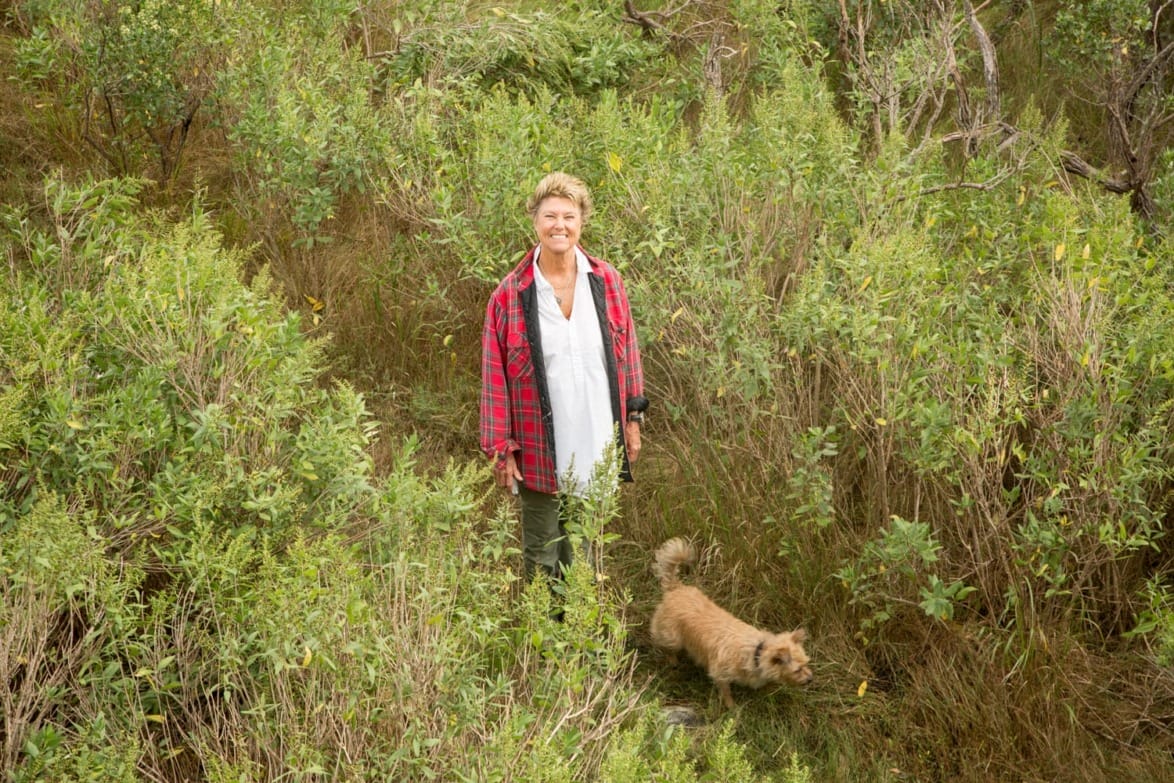 Woman in red plaid jacket stands smiling in tall grass with a small dog nearby among green bushes.