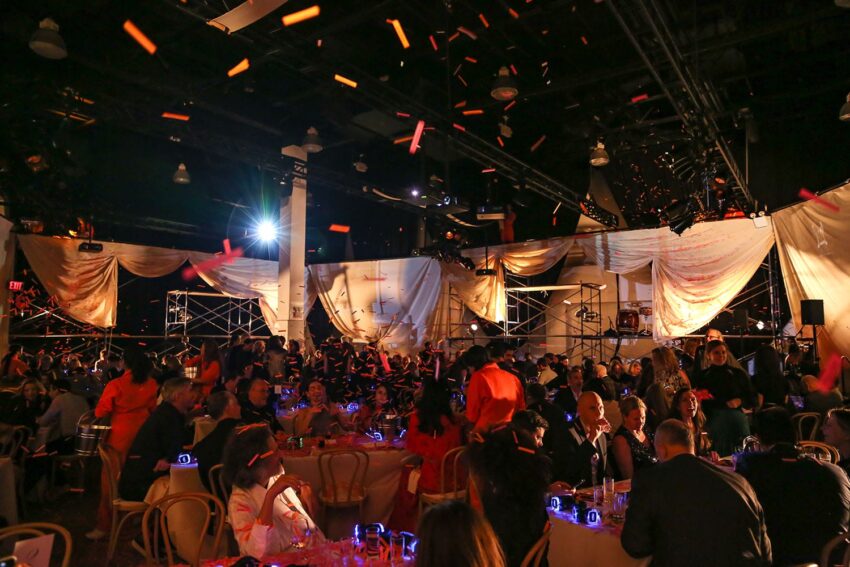 A lively, crowded event with people seated at tables, colorful decorations, and vibrant lighting in an indoor venue.
