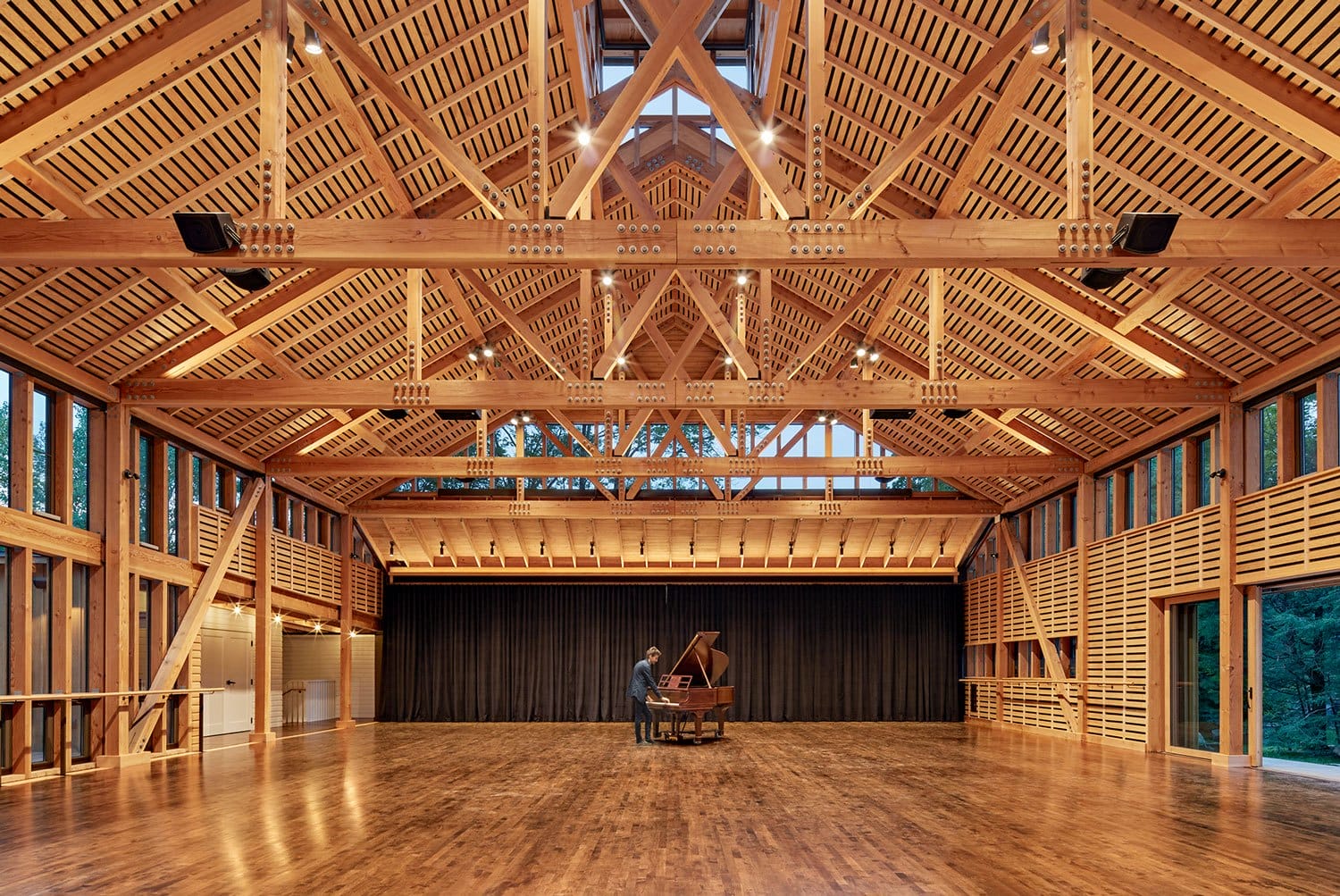 Spacious wooden hall with a grand piano and a person playing, surrounded by natural light from large windows.