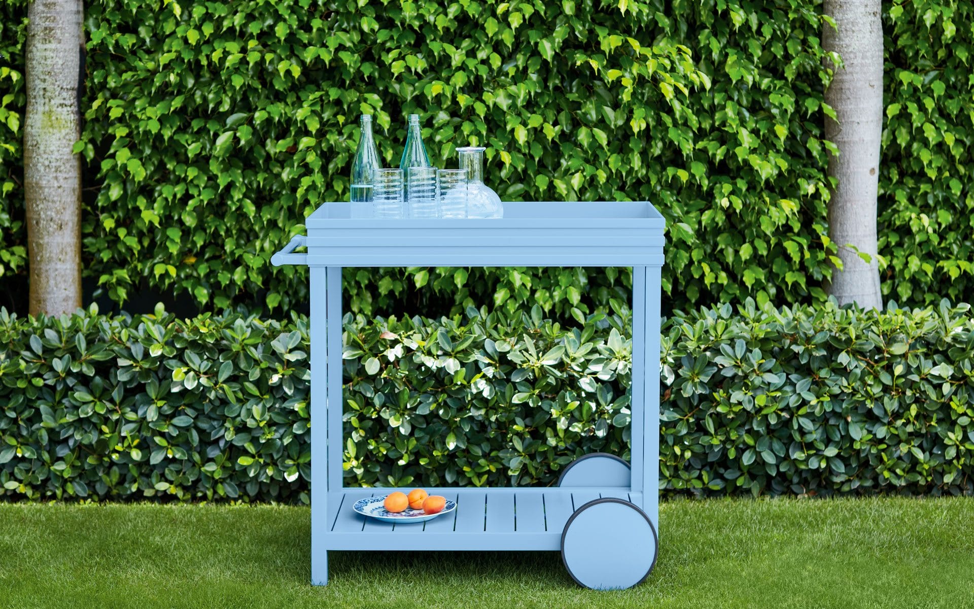 Blue outdoor bar cart with glasses and bottles on top, oranges on bottom shelf, set against a lush green hedge background.