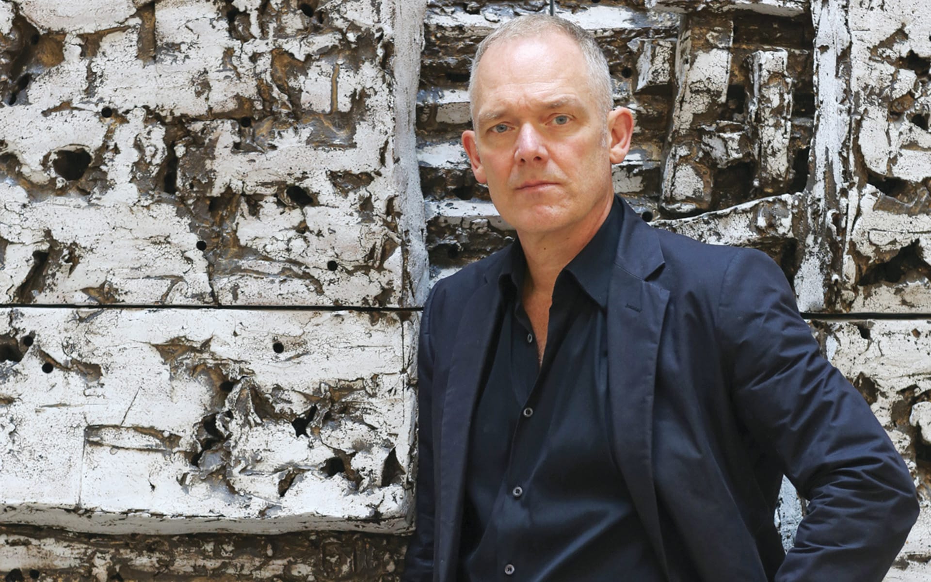 A person in a black jacket stands in front of a textured, abstract white wall.