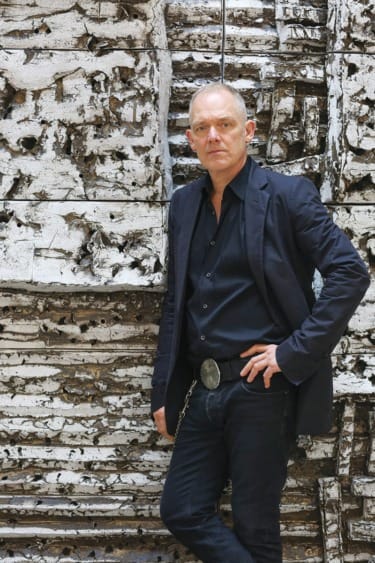 Person stands confidently in front of a textured, abstract background wearing a dark outfit and a belt with a large buckle.