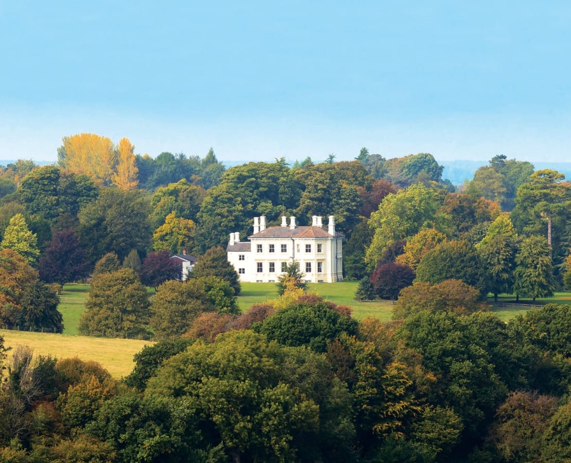 White mansion surrounded by lush greenery and colorful autumn trees under a clear blue sky.
