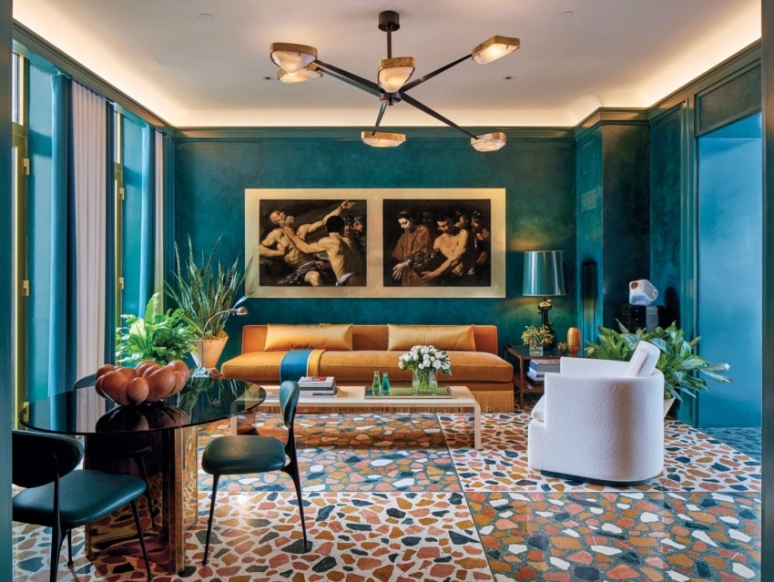 Modern living room with teal walls, colorful mosaic floor, orange sofa, round black table, and framed paintings on the wall.
