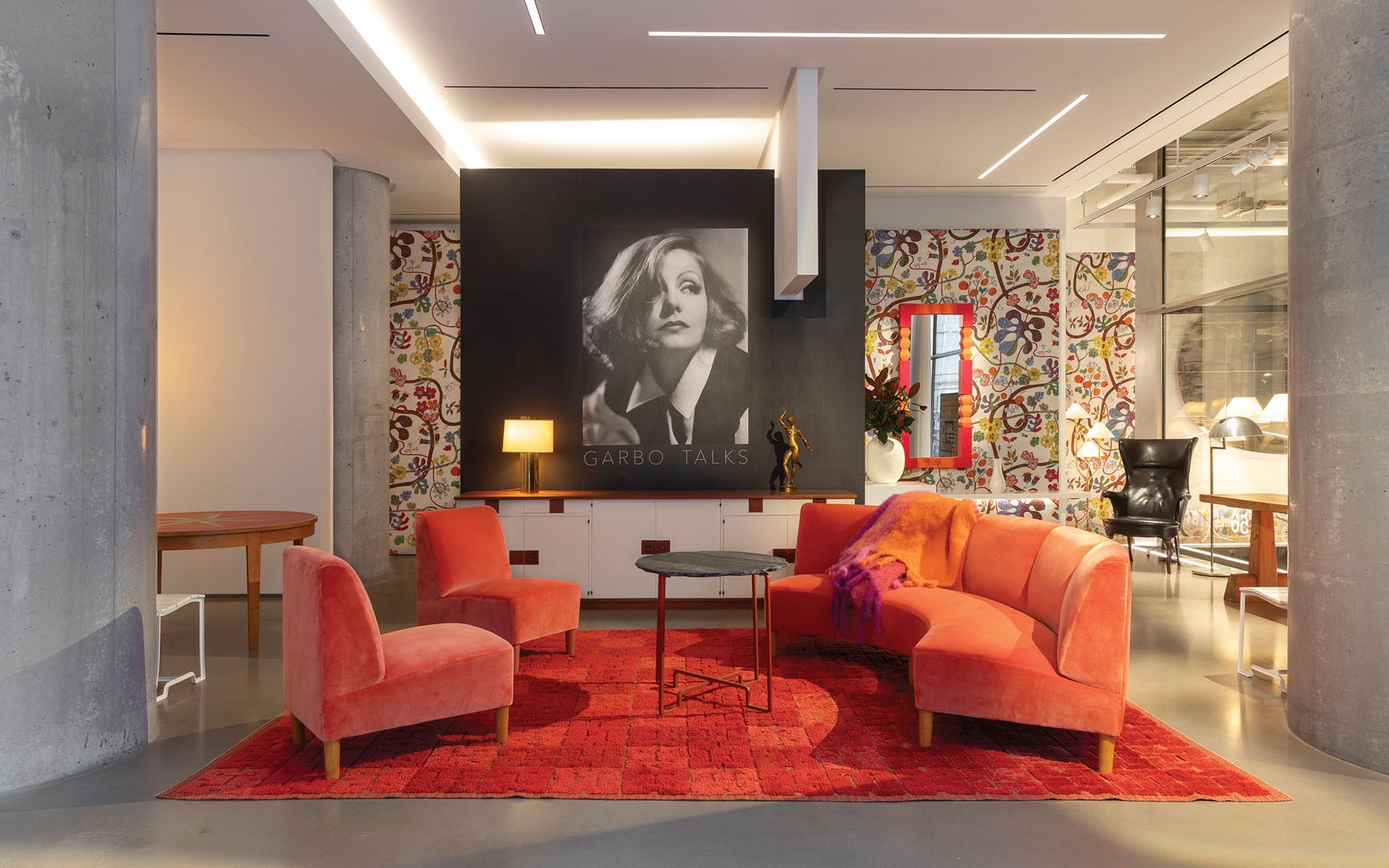 Modern living room with red sofas, round table, vibrant rug, and artistic black-and-white wall portrait.