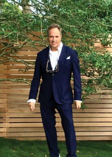 Man in navy suit standing in front of a wooden fence and greenery outdoors.