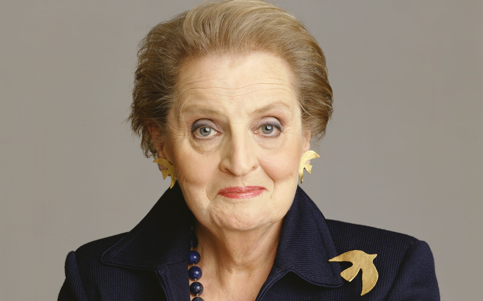 Elderly woman with short hair and a serious expression, wearing a dark jacket with bird-shaped pins and matching earrings.