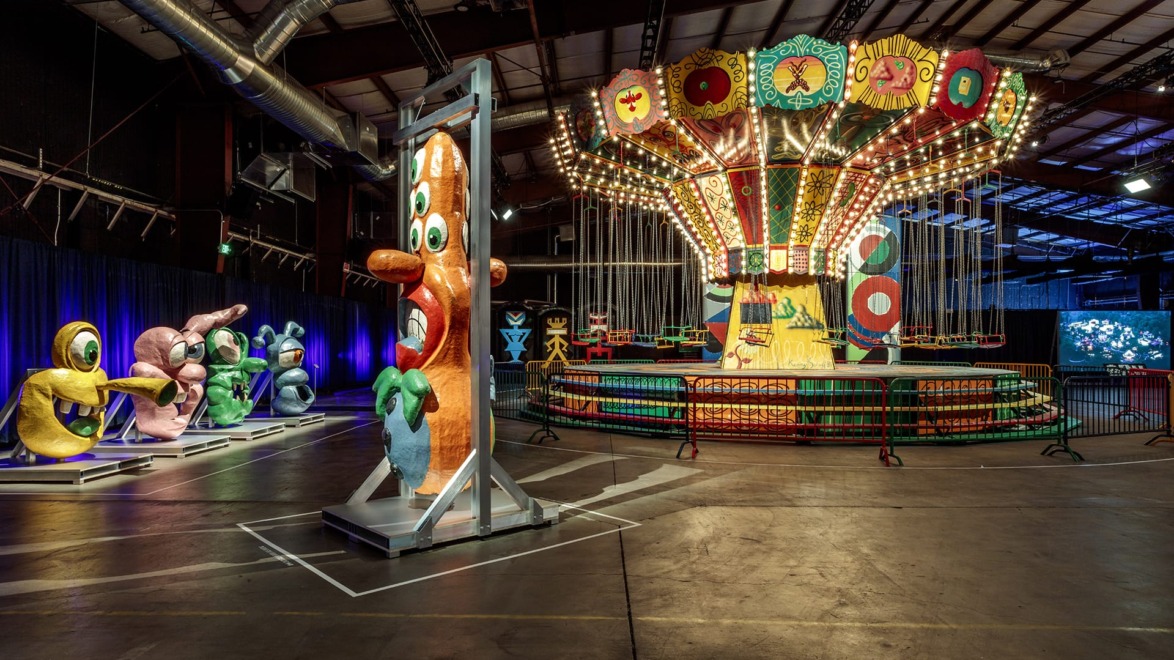 Colorful, whimsical carnival ride indoors with vibrant, abstract sculptures in a dimly lit warehouse setting.