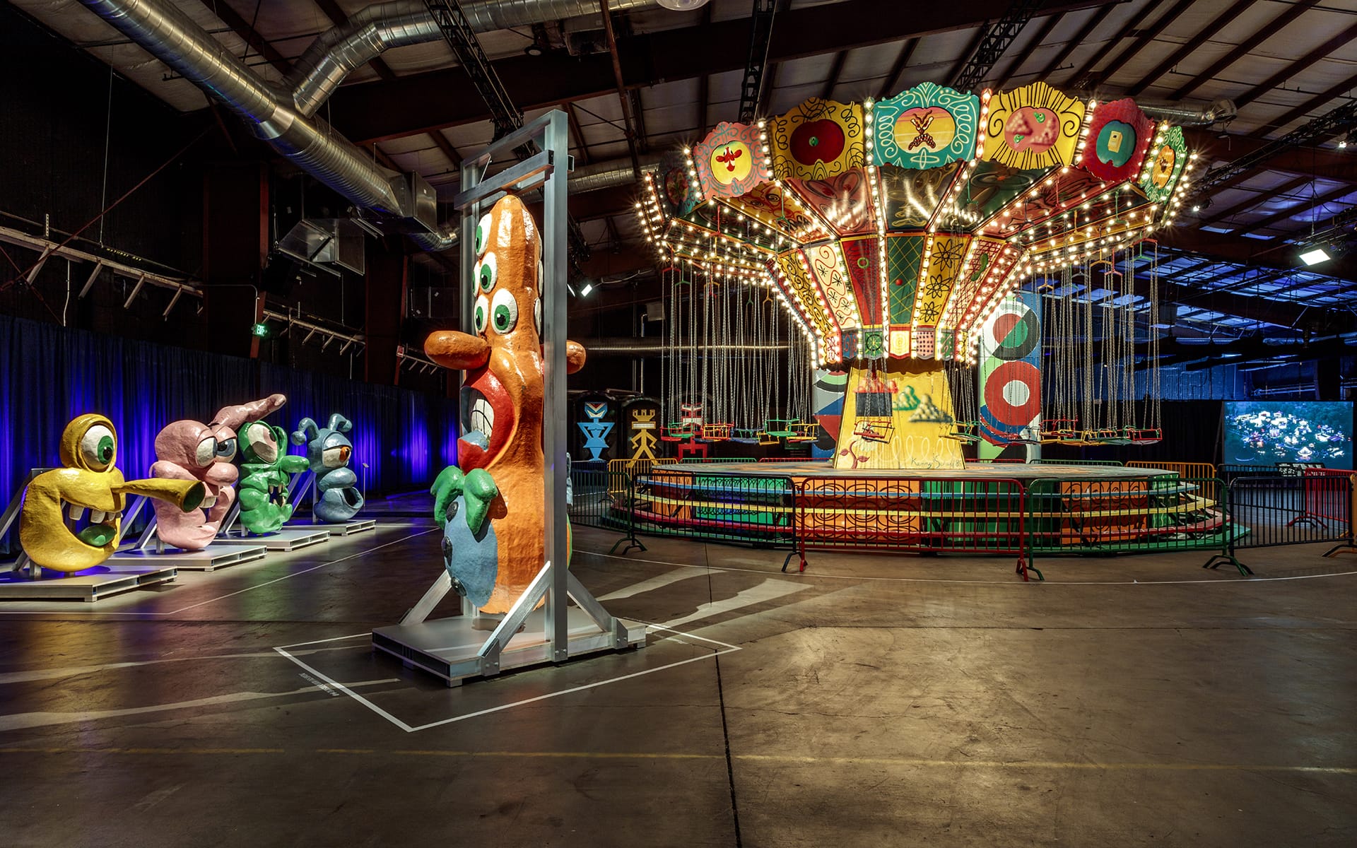 Colorful, whimsical carnival ride indoors with vibrant, abstract sculptures in a dimly lit warehouse setting.