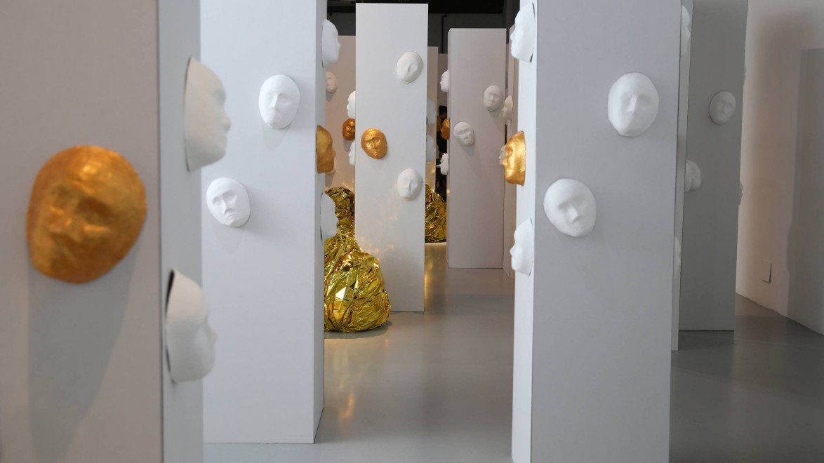 Art installation featuring white pillars adorned with white and gold face sculptures in a modern gallery setting.