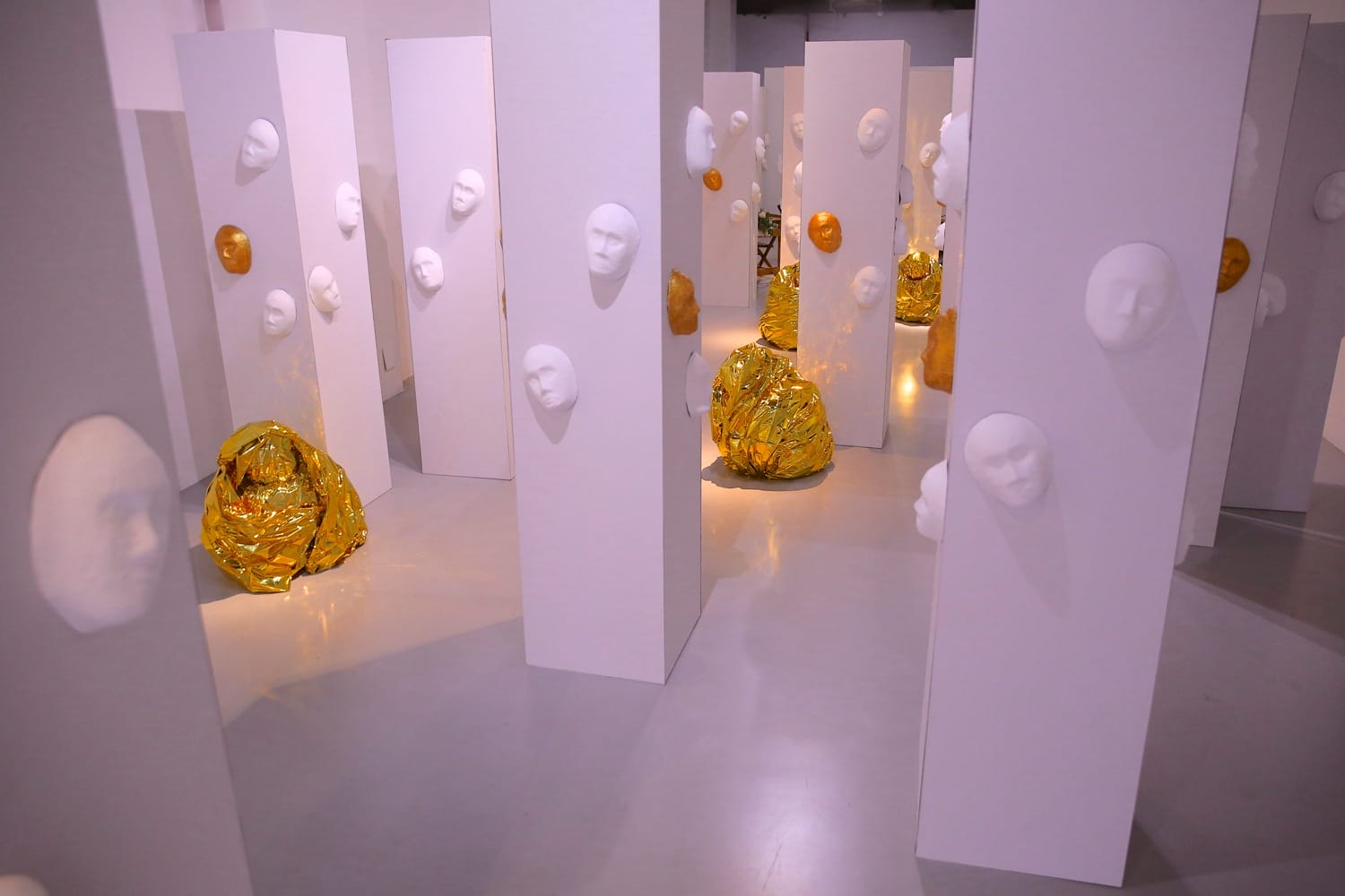 Art installation with white pillars, sculpted faces, and yellow crumpled material on the floor, creating an abstract scene.