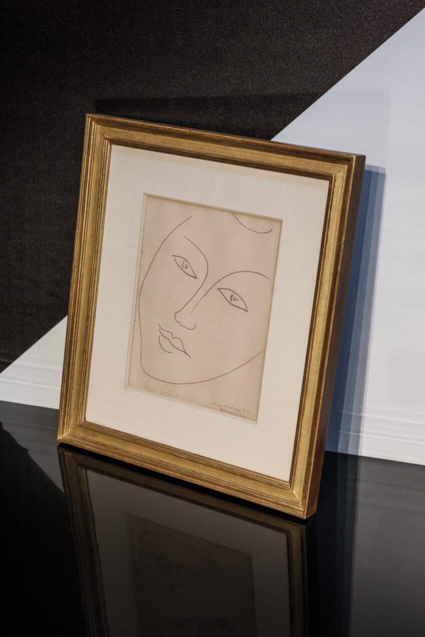 Framed minimalist sketch of a face with a gold border, displayed on a reflective black surface.