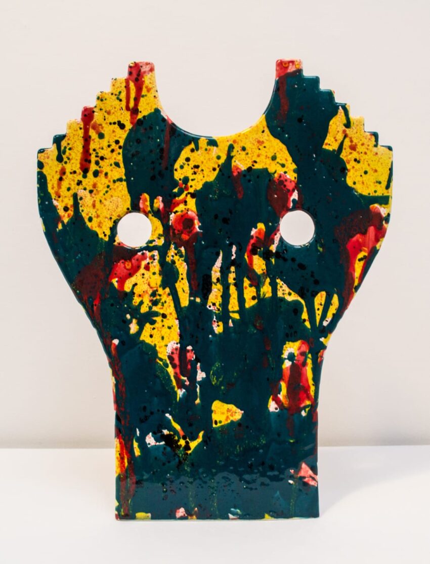 Abstract ceramic sculpture with vibrant yellow, red, and green splashes, featuring two circular cutouts on a white background.