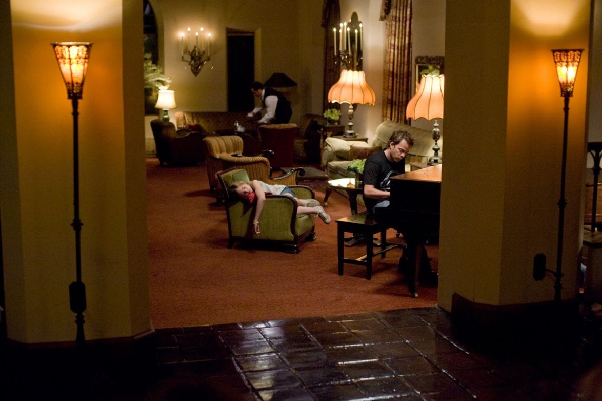 Man playing piano in a warmly lit lounge with vintage decor, another person reclining on a nearby chair, and a standing figure.