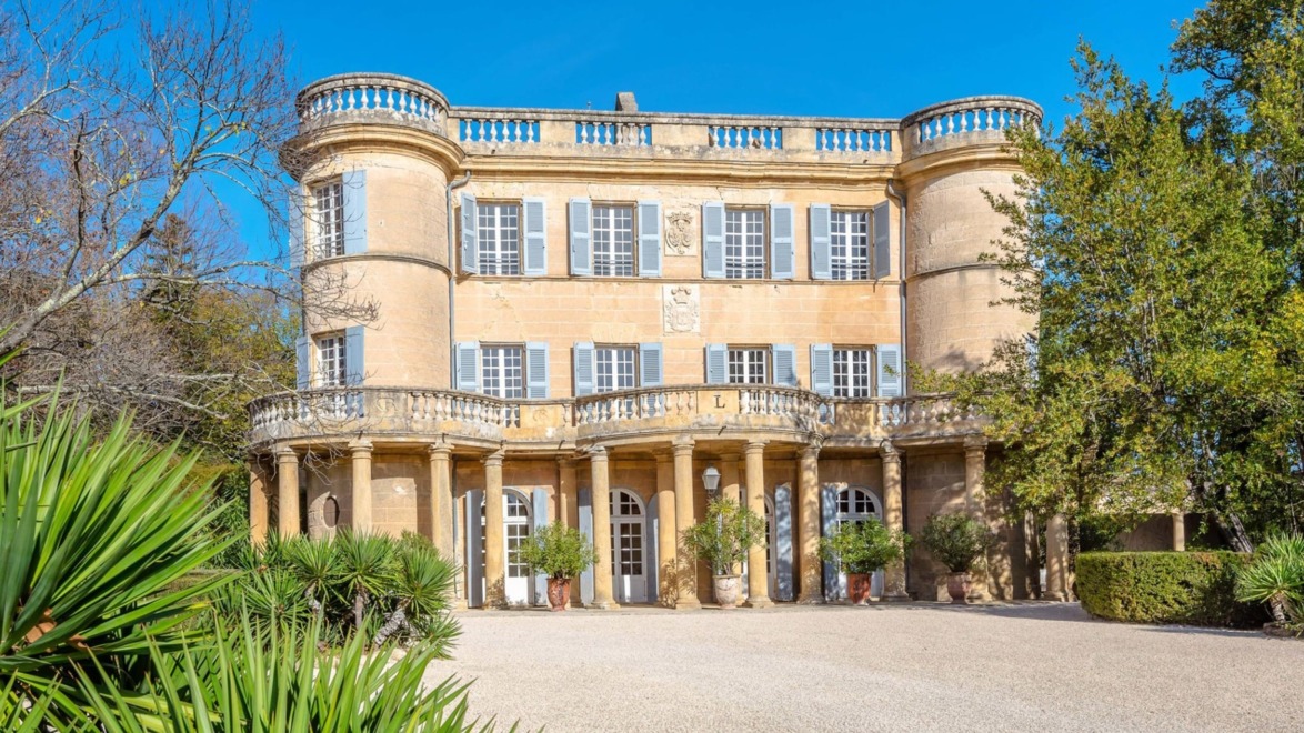 Historic mansion with round towers, large windows, and decorative balcony set in a lush garden under a clear blue sky.