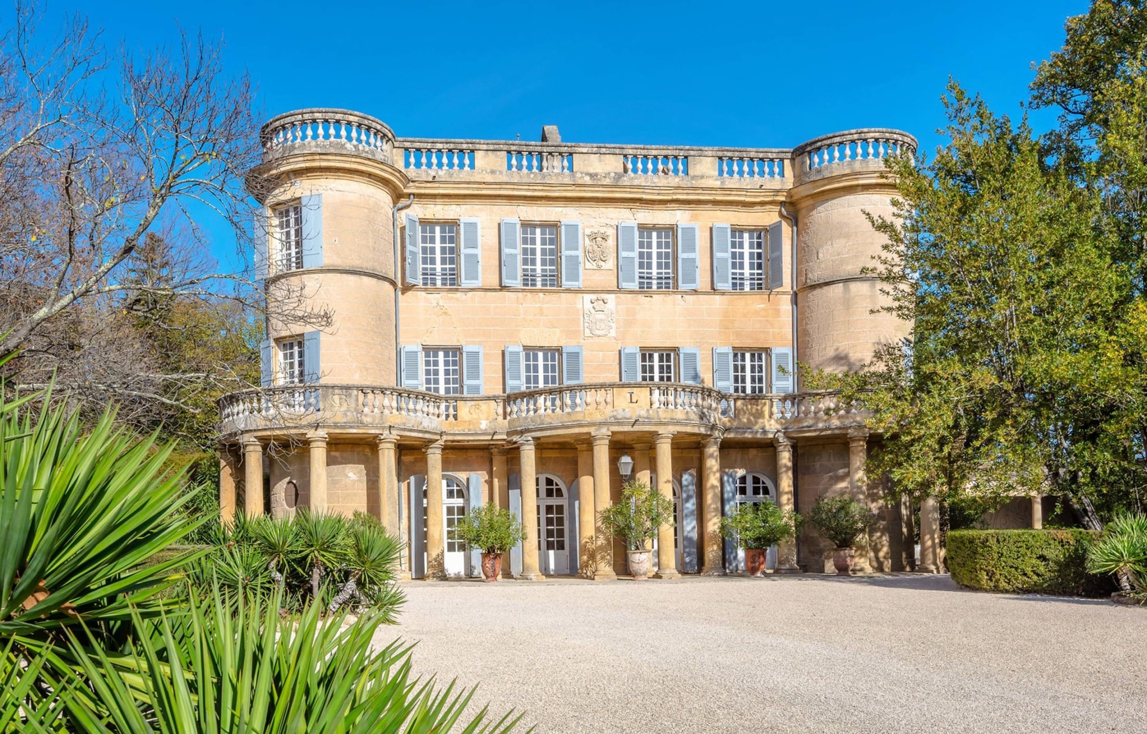 Historic mansion with round towers, large windows, and decorative balcony set in a lush garden under a clear blue sky.