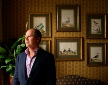 Man in a suit looking out a window, standing in a room with framed bird art and patterned wallpaper.