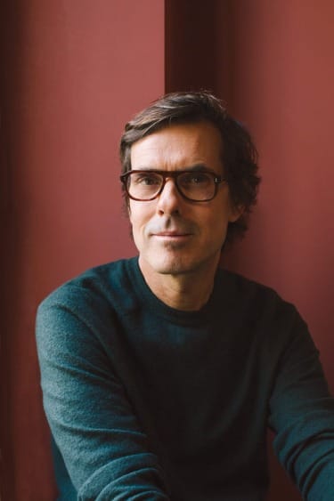 Man with glasses sitting against a red wall, wearing a dark sweater, looking at the camera.