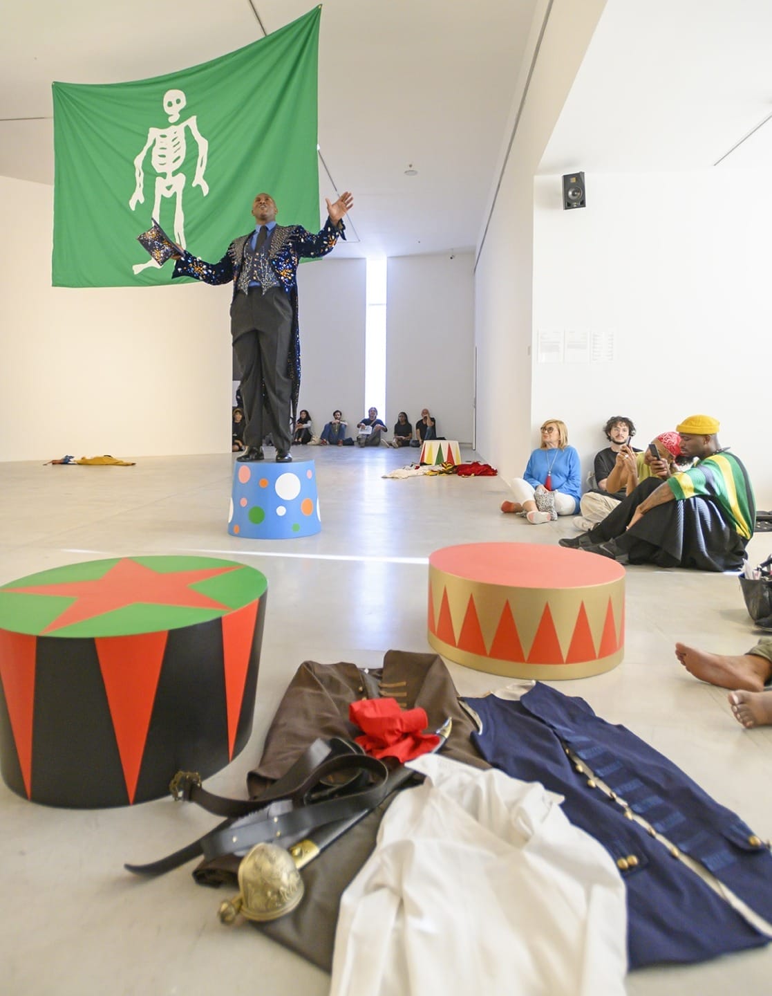 Performer stands on colorful pedestal with onlookers seated nearby in a modern art gallery, green flag with skeleton in background.