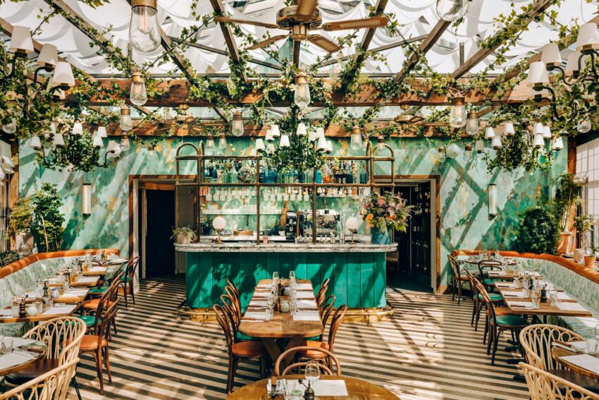Stylish, sunlit restaurant interior with green walls, wooden furniture, and hanging plants, centered around a teal bar.