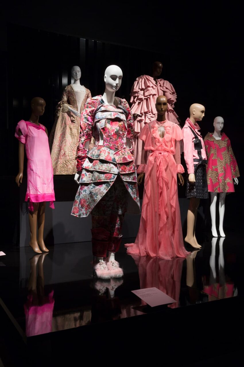 Fashion exhibit with mannequins displaying vibrant, layered clothing in pink and floral patterns on a dark backdrop.