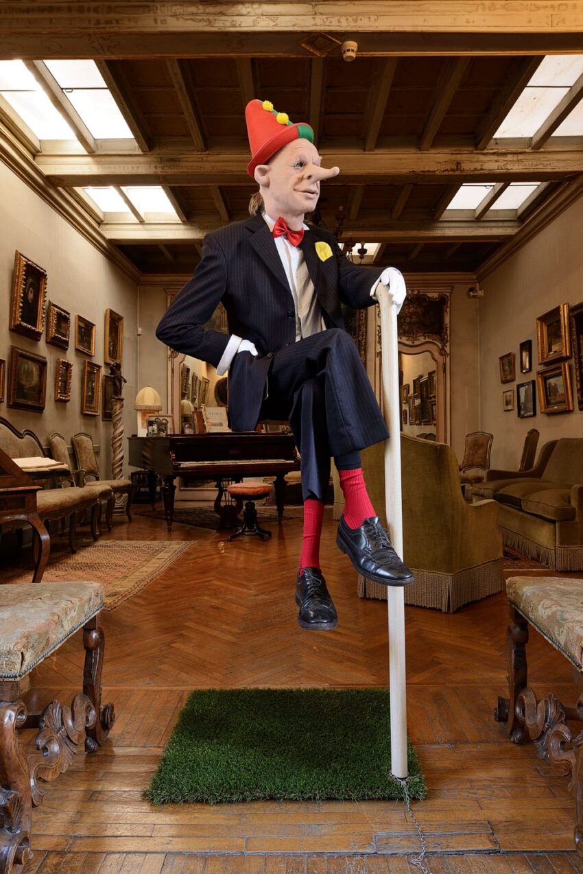 Man in a suit and clown hat balancing on a pole in an elegant room with wooden floors and paintings on the walls.