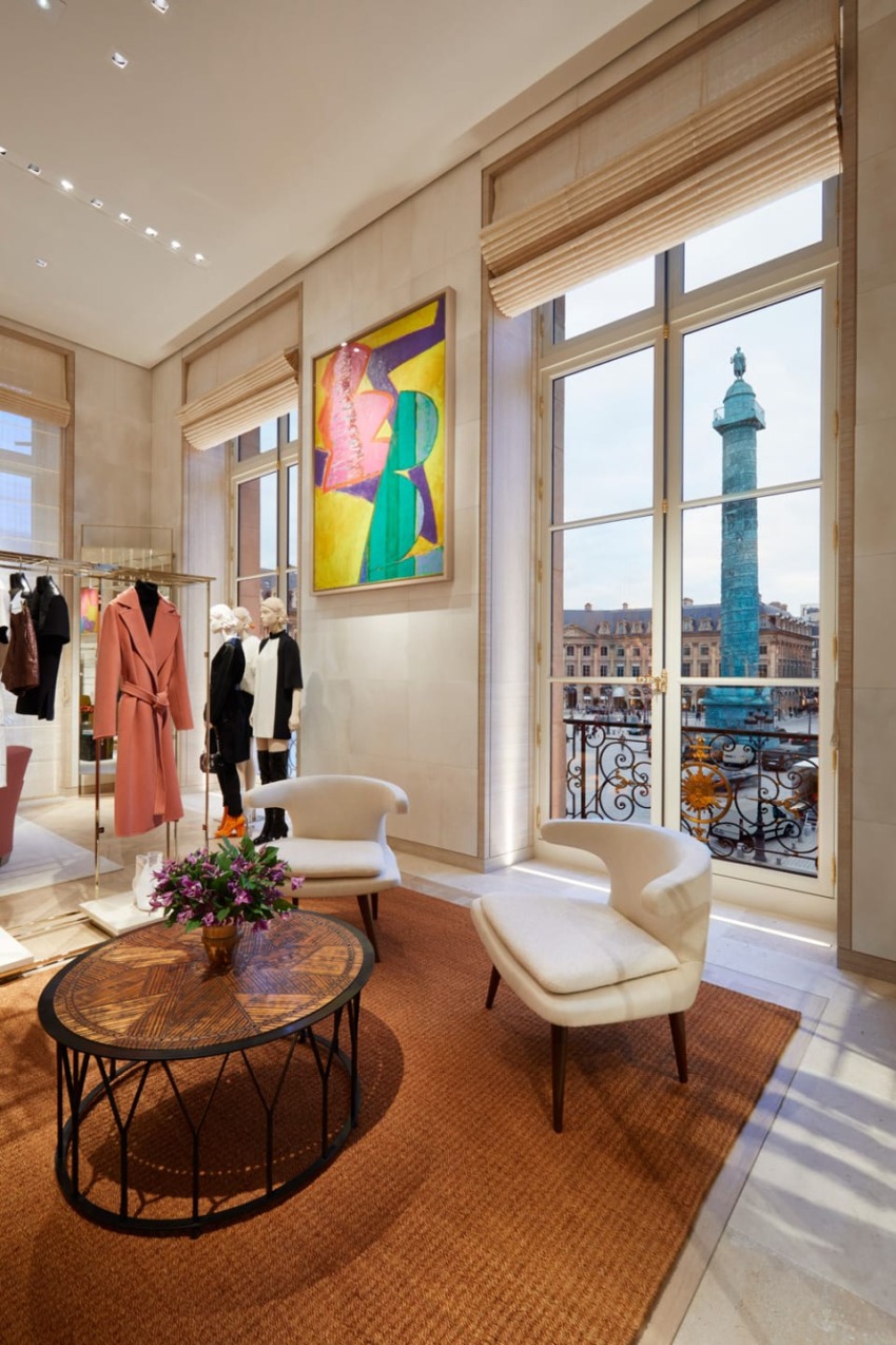 Luxury clothing store interior with modern art, stylish chairs, and a view of a city square through large windows.