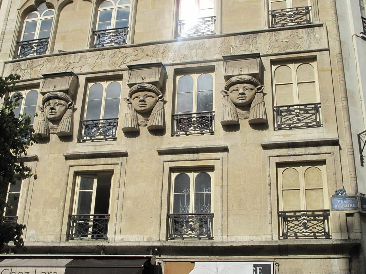 Facade of a building with three stone face sculptures and decorative metal railings on windows.
