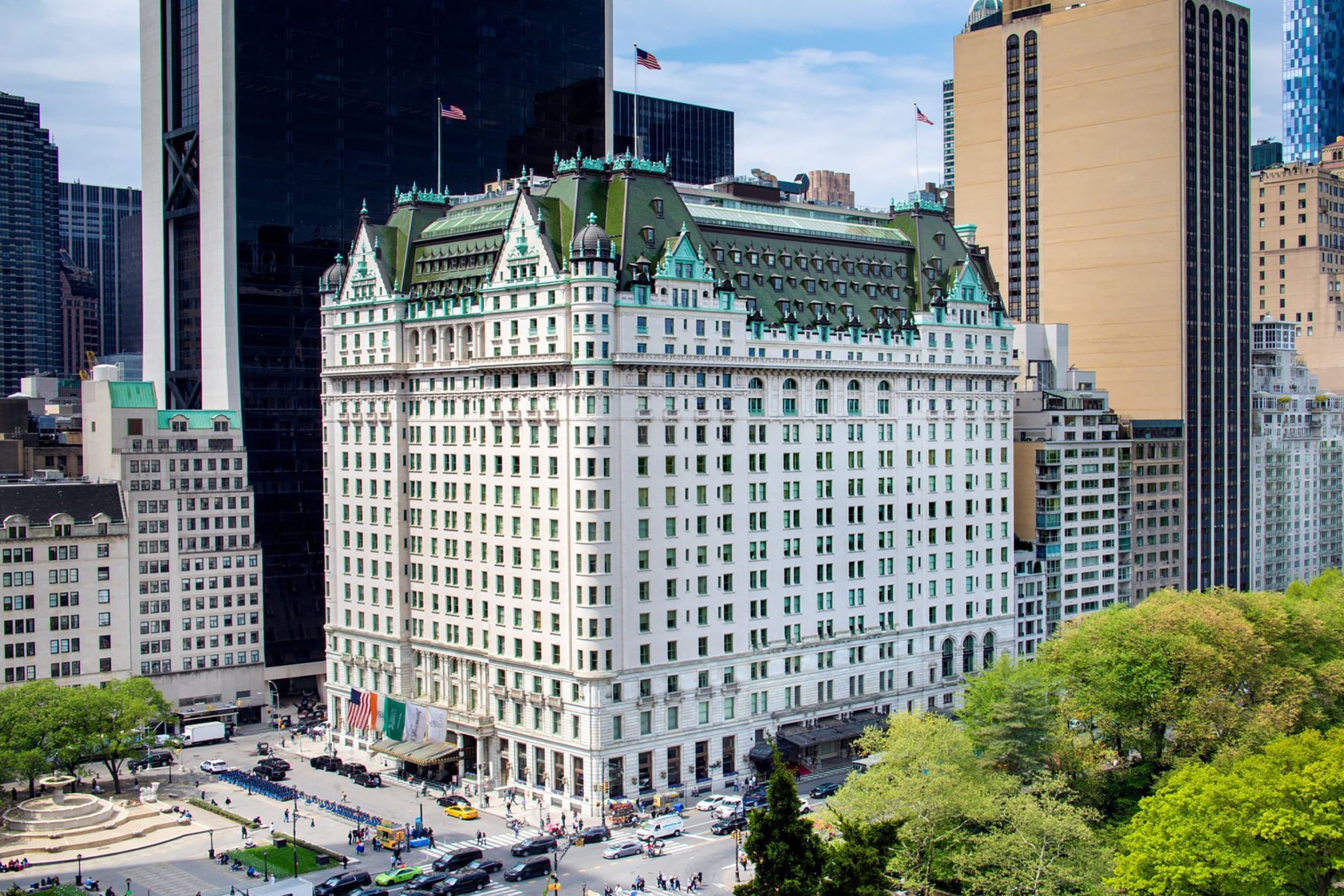 Aerial view of a large, historic hotel building with green roof, surrounded by skyscrapers and trees in an urban setting.