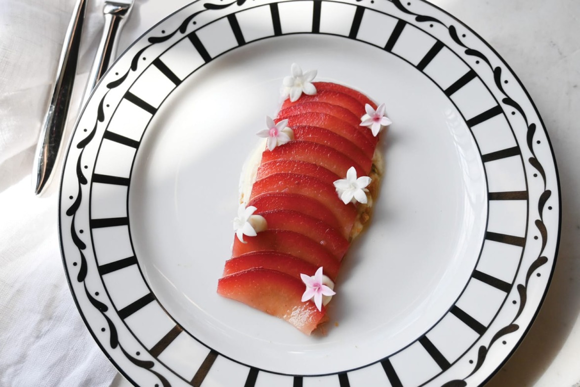 Sliced red fruit arranged on a decorative plate, garnished with small white flowers, next to silver cutlery on a white tablecloth.