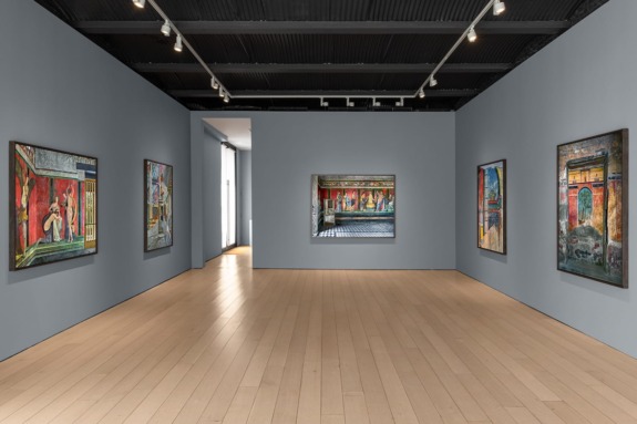 Art gallery interior showcasing colorful paintings on gray walls with a wooden floor and track lighting above.