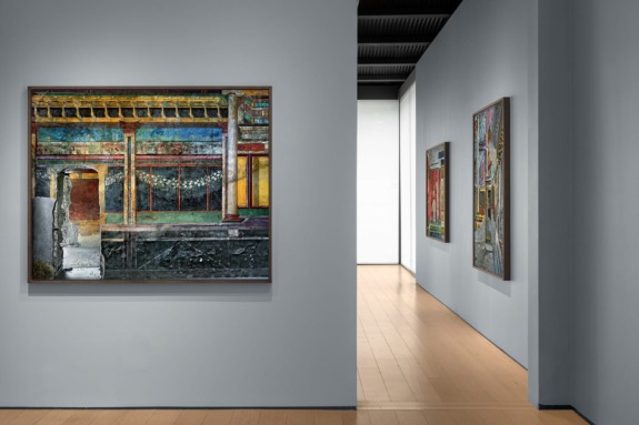 Art gallery with modern paintings on blue-grey walls and wooden floor, featuring vibrant abstract and architectural scenes.