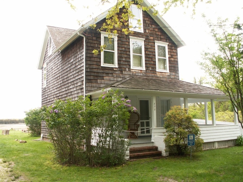 Two-story house with shingle siding, front porch, and green lawn surrounded by trees and shrubs.
