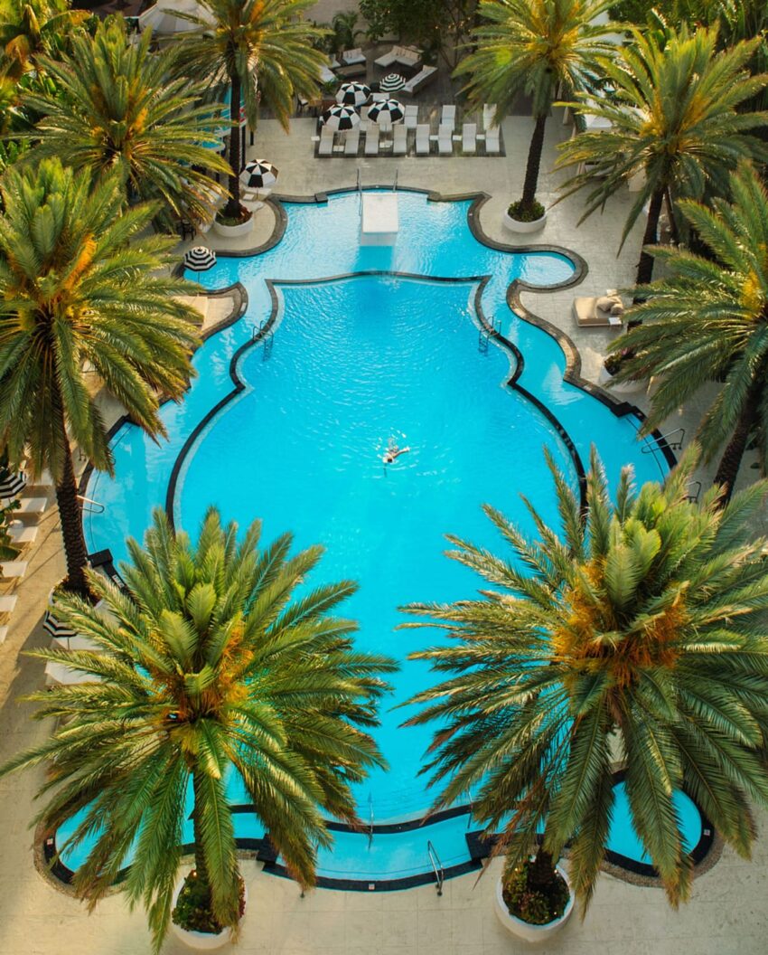 Aerial view of a luxurious hotel pool surrounded by palm trees and sun loungers with people swimming and relaxing.