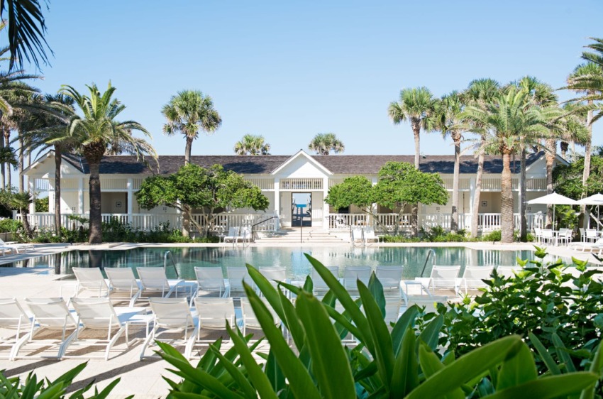 Luxurious resort pool area with palm trees and white lounge chairs in front of a large white building under a clear blue sky