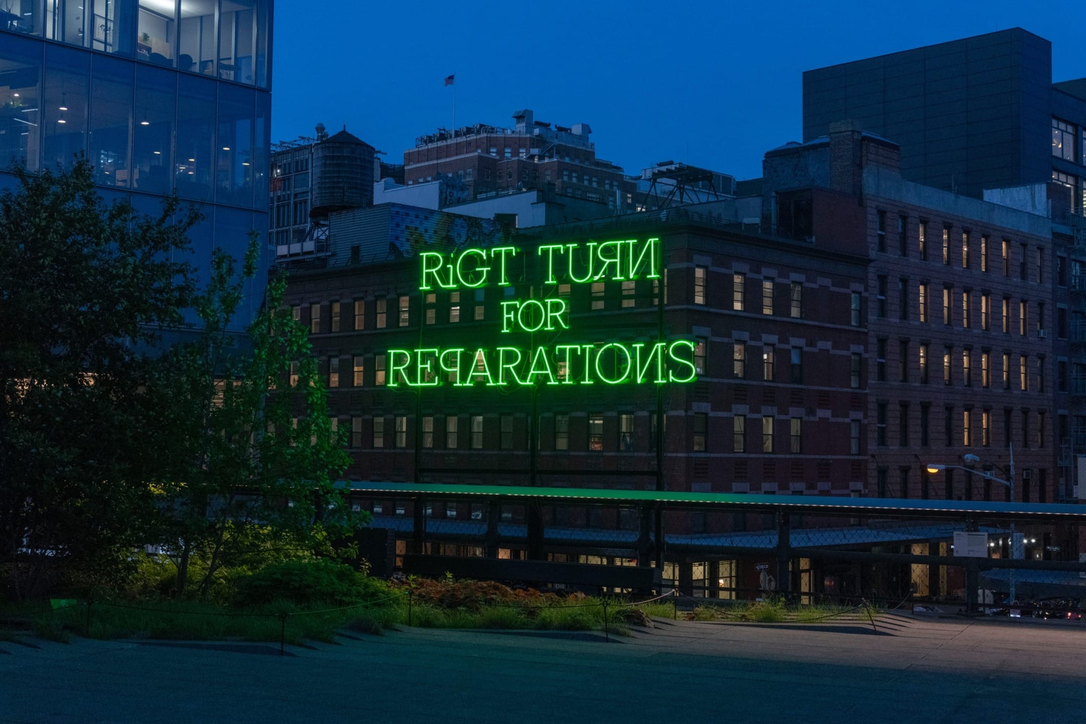 Neon sign reading "Right Turn for Reparations" on a brick building at night with cityscape in the background.