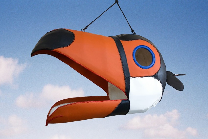 Large orange bird sculpture hanging against a blue sky with clouds, resembling a stylized toucan head.