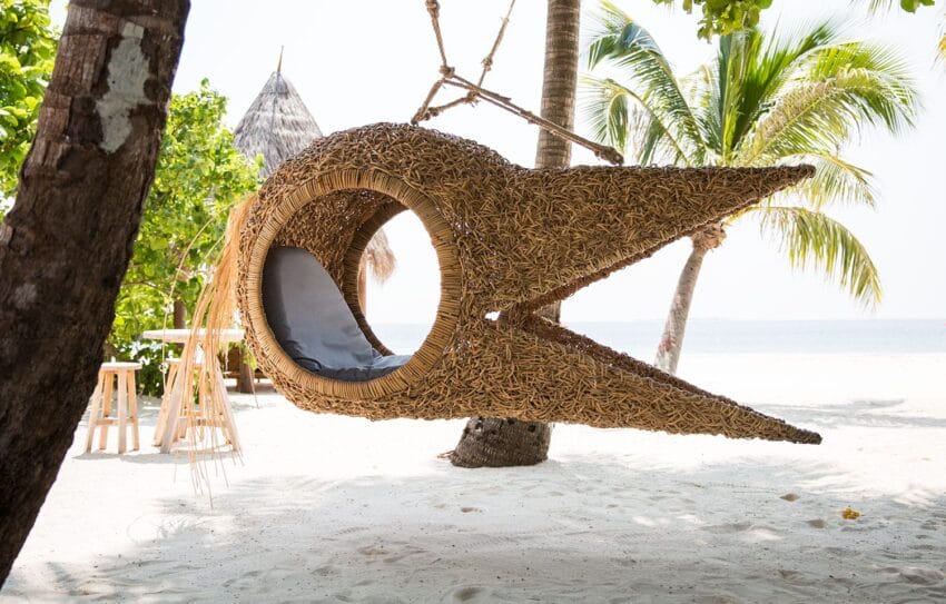 Wicker pod hanging from a tree on a sandy beach, with palm trees and ocean in the background.