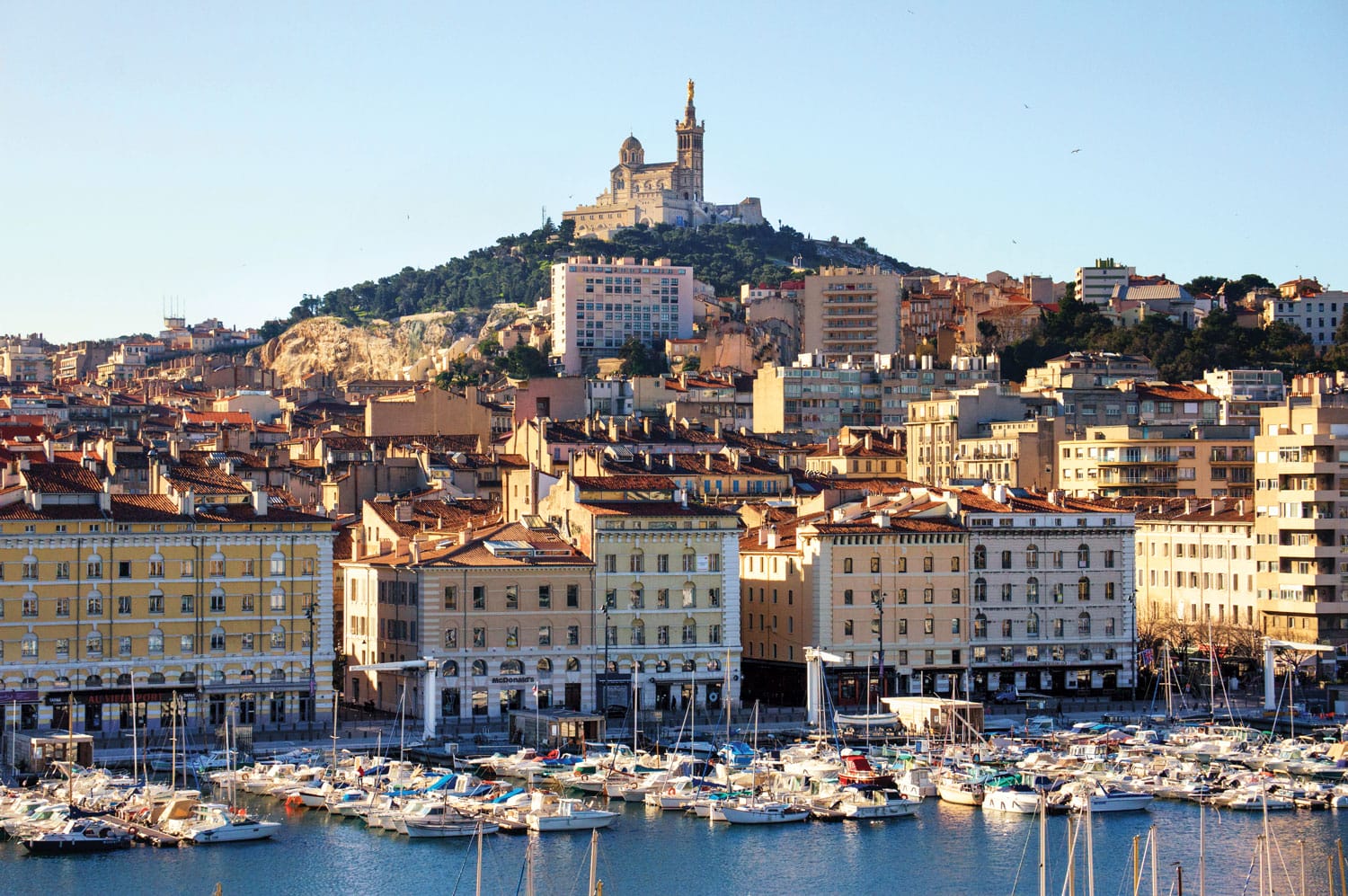 Scenic view of Marseille port with boats and historic buildings under a clear sky, featuring a hilltop basilica in the background.