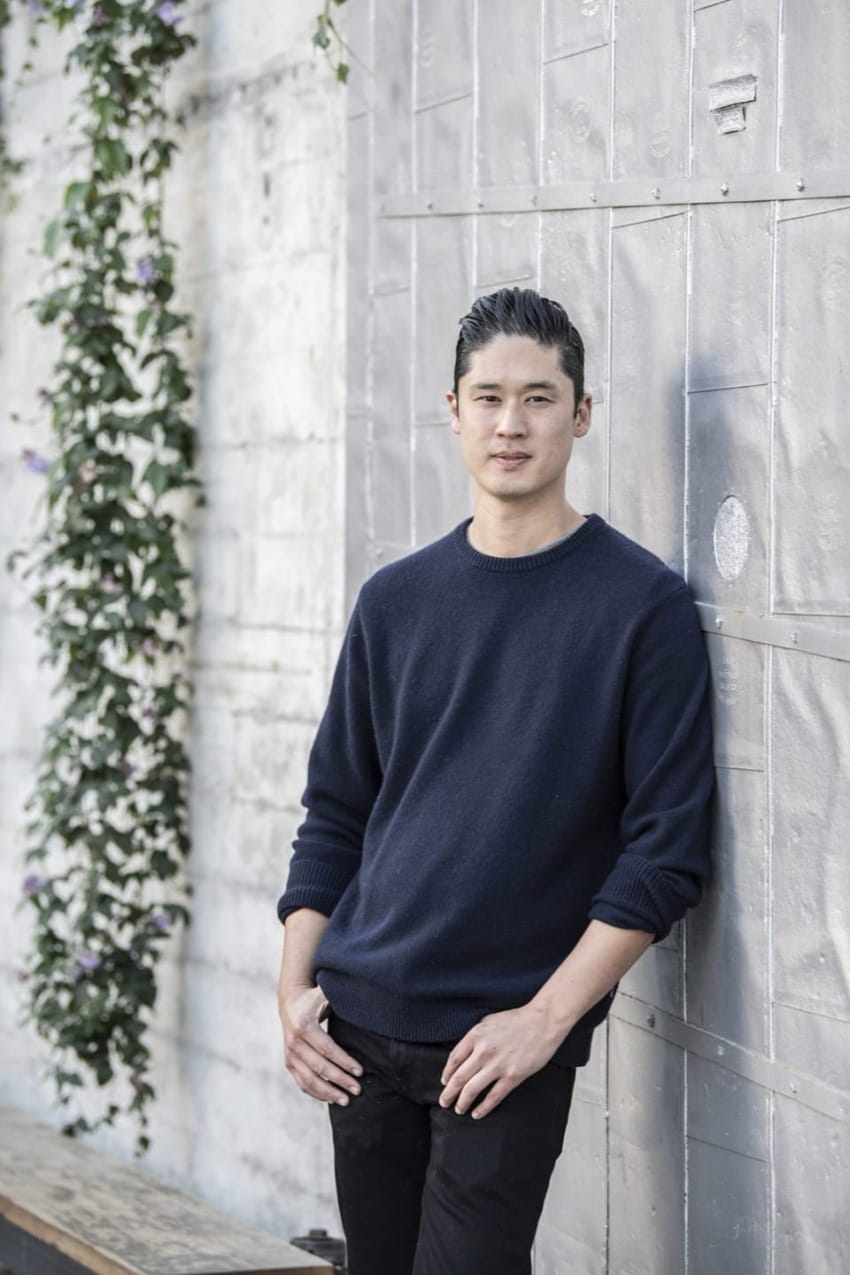 Man in dark sweater leaning against a textured wall, with plants climbing nearby.